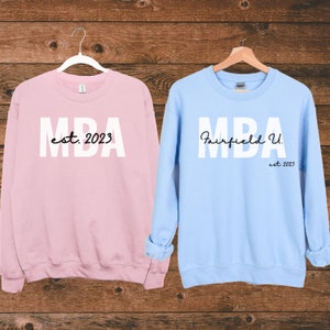 May include: Two sweatshirts, one pink and one light blue, with the text "MBA est. 2023" on the pink sweatshirt and "MBA Fairfield U. est. 2023" on the light blue sweatshirt.