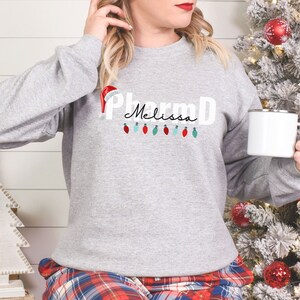 May include: A gray sweatshirt with the text "PharmD Melissa" and a Santa hat on top of the "P".  There are red and green Christmas lights hanging below the text.