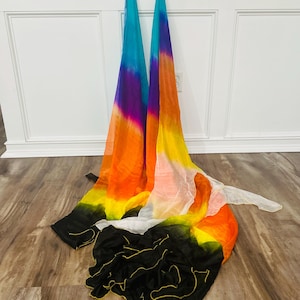 May include: Two colorful silk dance veils with a gradient of colors, including blue, purple, orange, yellow, and black. The veils are draped on a wooden floor, with a white wall in the background. The veils are used for dance and performance.