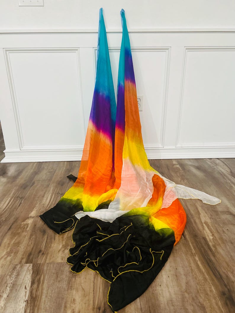 May include: Two colorful silk dance veils with a gradient of colors, including blue, purple, orange, yellow, and black. The veils are draped on a wooden floor, with a white wall in the background. The veils are used for dance and performance.