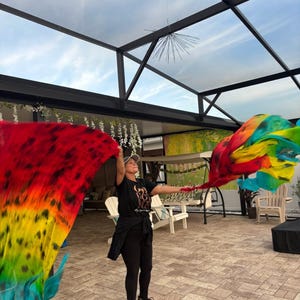 May include: A person holding up two large, colorful fabric flags. The flags display a vibrant gradient of red, orange, yellow, green, and blue. The person is wearing a black t-shirt, leggings, and a cap.