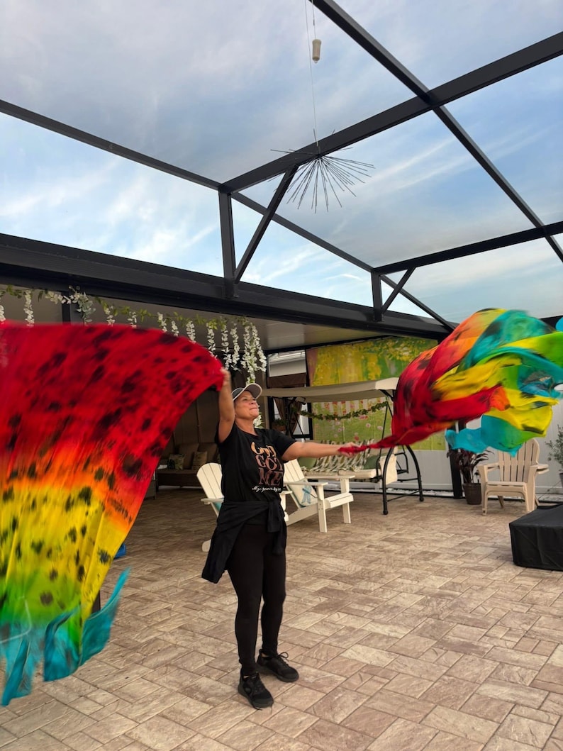 May include: A person holding up two large, colorful fabric flags. The flags display a vibrant gradient of red, orange, yellow, green, and blue. The person is wearing a black t-shirt, leggings, and a cap.