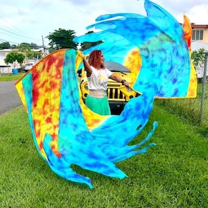 May include: A person is holding and waving a large, colorful flag outdoors. The flag features vibrant hues of blue, yellow, orange, and red. The person is wearing a white top and green skirt. A yellow vehicle is visible in the background, along with green grass and a cloudy sky.
