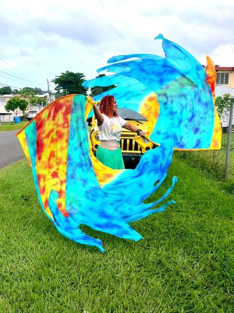 May include: A person is holding and waving a large, colorful flag outdoors. The flag features vibrant hues of blue, yellow, orange, and red. The person is wearing a white top and green skirt. A yellow vehicle is visible in the background, along with green grass and a cloudy sky.