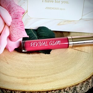 May include: A pink and gold tube of lip gloss with the words "Revival Glam" printed on the tube. The tube is sitting on a wooden surface. The background includes a pink rose and a card with the text "For I know the plans I have for you. Jeremiah 29:11"