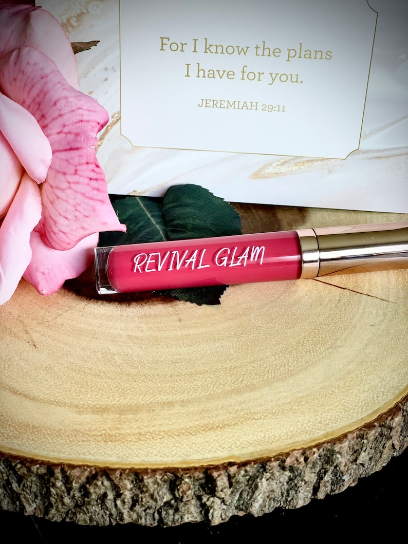 May include: A pink and gold tube of lip gloss with the words "Revival Glam" printed on the tube. The tube is sitting on a wooden surface. The background includes a pink rose and a card with the text "For I know the plans I have for you. Jeremiah 29:11"