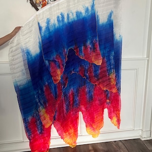 May include: A flowing, colorful fabric with a tie-dye effect. The top portion is white, transitioning to blue, red, and yellow hues. The fabric is held up, showcasing its vibrant design and texture. The overall theme is artistic and decorative.