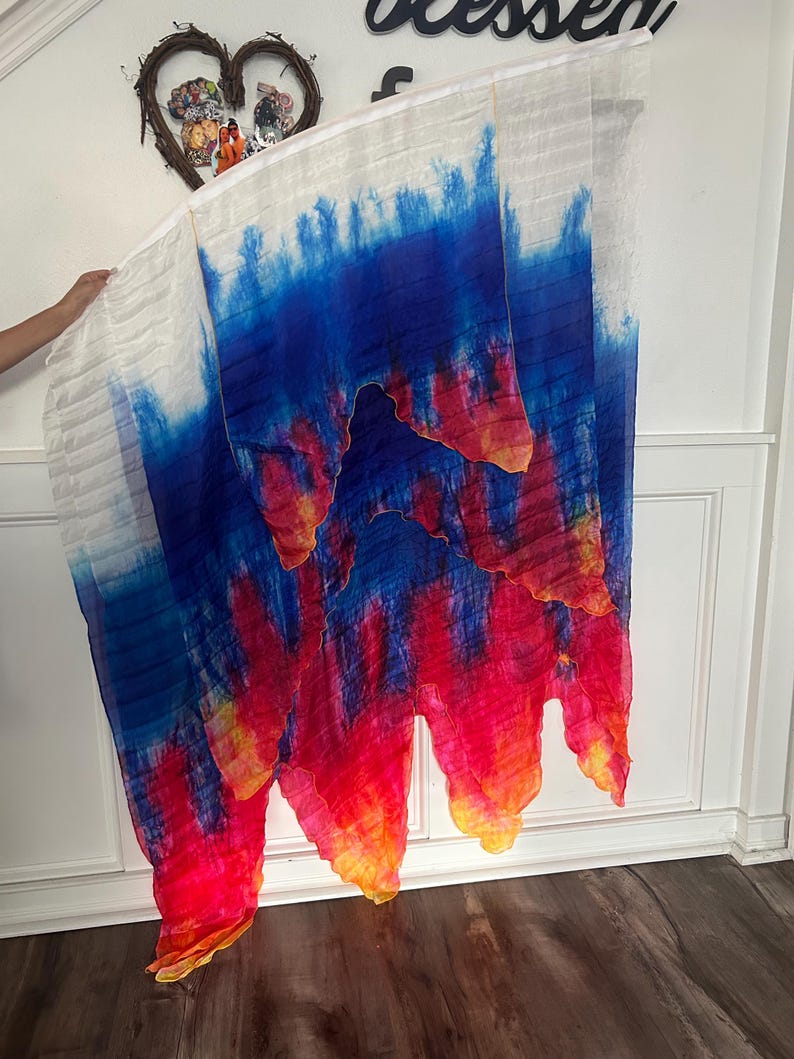 May include: A flowing, colorful fabric with a tie-dye effect. The top portion is white, transitioning to blue, red, and yellow hues. The fabric is held up, showcasing its vibrant design and texture. The overall theme is artistic and decorative.