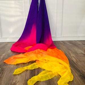 May include: A flowing, colorful fabric in a gradient of colors, from purple to pink, orange, and yellow. The fabric is draped on a wooden floor, with the top portion hanging against a white wall. The fabric's vibrant colors create a visually striking display.