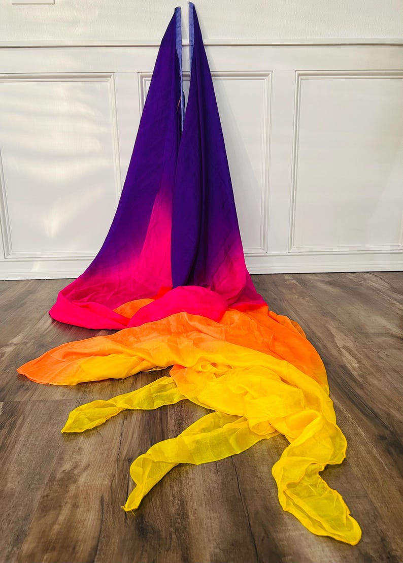 May include: A flowing, colorful fabric in a gradient of colors, from purple to pink, orange, and yellow. The fabric is draped on a wooden floor, with the top portion hanging against a white wall. The fabric's vibrant colors create a visually striking display.
