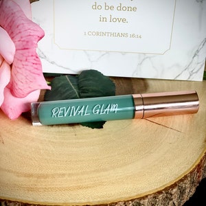 May include: A green tube of lip gloss with a rose gold cap. The tube has the words "REVIVAL GLAM" printed on it. The background is a wooden surface with a pink rose.