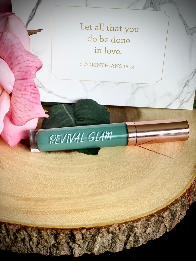 May include: A green tube of lip gloss with a rose gold cap. The tube has the words "REVIVAL GLAM" printed on it. The background is a wooden surface with a pink rose.
