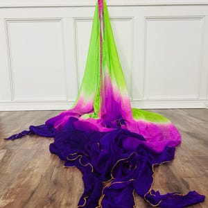May include: A flowing, multi-colored fabric in shades of green, pink, and purple. The fabric is draped across a wooden floor. The edges of the purple fabric are trimmed with gold. The fabric is likely used for dance or performance.