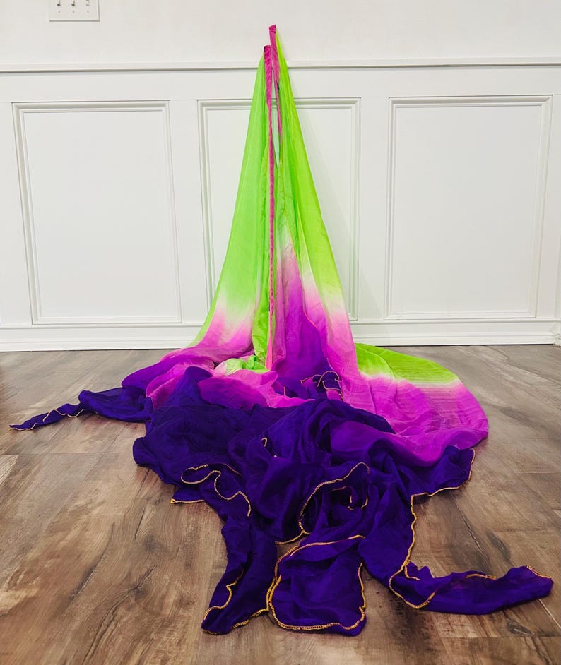 May include: A flowing, multi-colored fabric in shades of green, pink, and purple. The fabric is draped across a wooden floor. The edges of the purple fabric are trimmed with gold. The fabric is likely used for dance or performance.