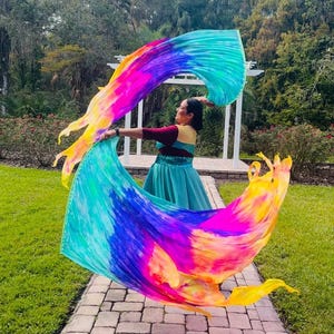 May include: A person is holding a large, colorful silk fan with vibrant hues of yellow, pink, blue, and purple. The fan is flowing in the air, creating a dynamic and artistic display. The person is wearing a teal dress and a maroon top.