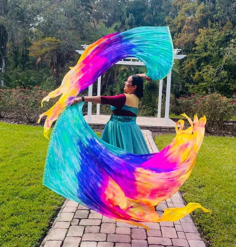 May include: A person is holding a large, colorful silk fan with vibrant hues of yellow, pink, blue, and purple. The fan is flowing in the air, creating a dynamic and artistic display. The person is wearing a teal dress and a maroon top.
