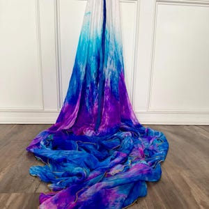 May include: A long, flowing scarf featuring a gradient tie-dye pattern that transitions from white at the top to vibrant shades of turquoise, royal blue, and deep purple at the bottom. The fabric has a lightweight, sheer texture with finished edges.