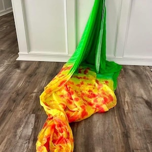 May include: A vibrant, flowing scarf in shades of green, yellow, and red. The scarf is draped on a wooden floor, with the colors blending in a dynamic pattern. The fabric appears lightweight and airy.
