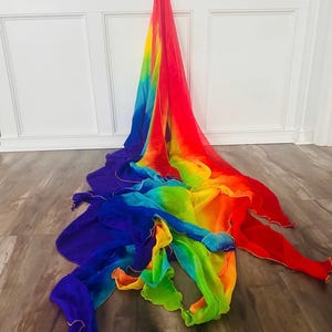 May include: A vibrant, rainbow-colored fabric draped on a wooden floor. The fabric transitions from red to orange, yellow, green, blue, and purple. The edges are trimmed with a gold-colored material. The fabric is likely used for dance or performance.