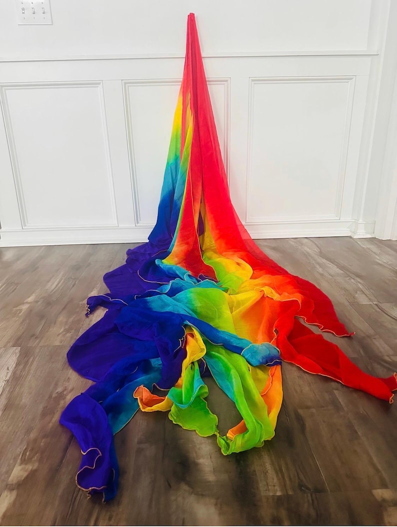 May include: A vibrant, rainbow-colored fabric draped on a wooden floor. The fabric transitions from red to orange, yellow, green, blue, and purple. The edges are trimmed with a gold-colored material. The fabric is likely used for dance or performance.