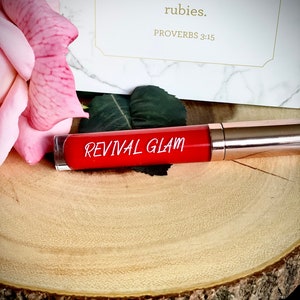 May include: A red tube of lip gloss with the words "Revival Glam" printed on the label. The tube is sitting on a wooden surface. The quote "She is more precious than rubies. Proverbs 3:15" is visible in the background.