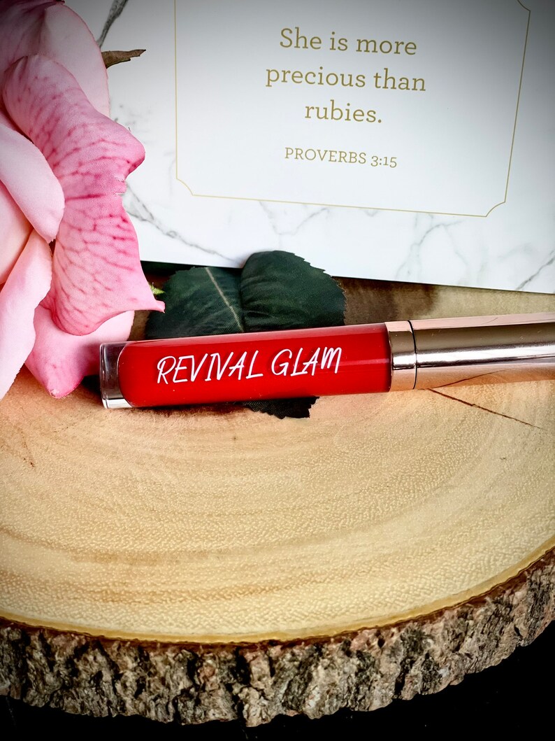 May include: A red tube of lip gloss with the words "Revival Glam" printed on the label. The tube is sitting on a wooden surface. The quote "She is more precious than rubies. Proverbs 3:15" is visible in the background.