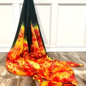 May include: A tie-dye scarf in shades of black, green, yellow, and red. The scarf is draped against a white wall and wooden floor. The fabric has a flowing, textured appearance, with the colors blending together.