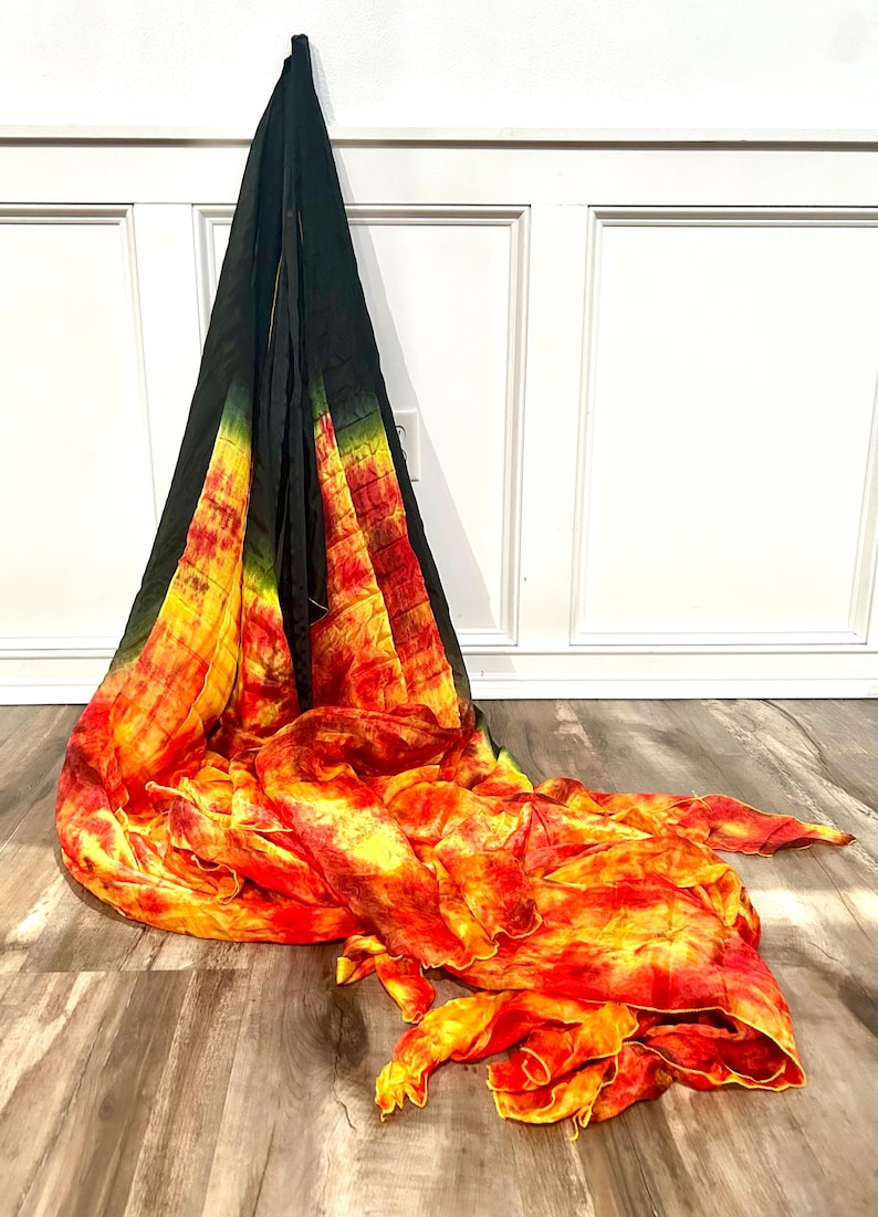 May include: A tie-dye scarf in shades of black, green, yellow, and red. The scarf is draped against a white wall and wooden floor. The fabric has a flowing, textured appearance, with the colors blending together.