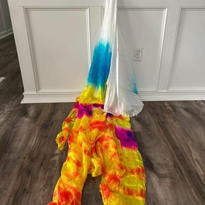 May include: A colorful, tie-dye fabric prop, likely for dance or performance. The fabric transitions from white to blue, yellow, orange, and pink. The fabric is draped against a white wall and a wooden floor.