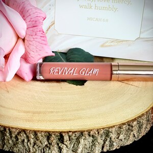 May include: A pink and nude lip gloss tube with the words "REVIVAL GLAM" printed on the label. The tube is sitting on a wooden surface.