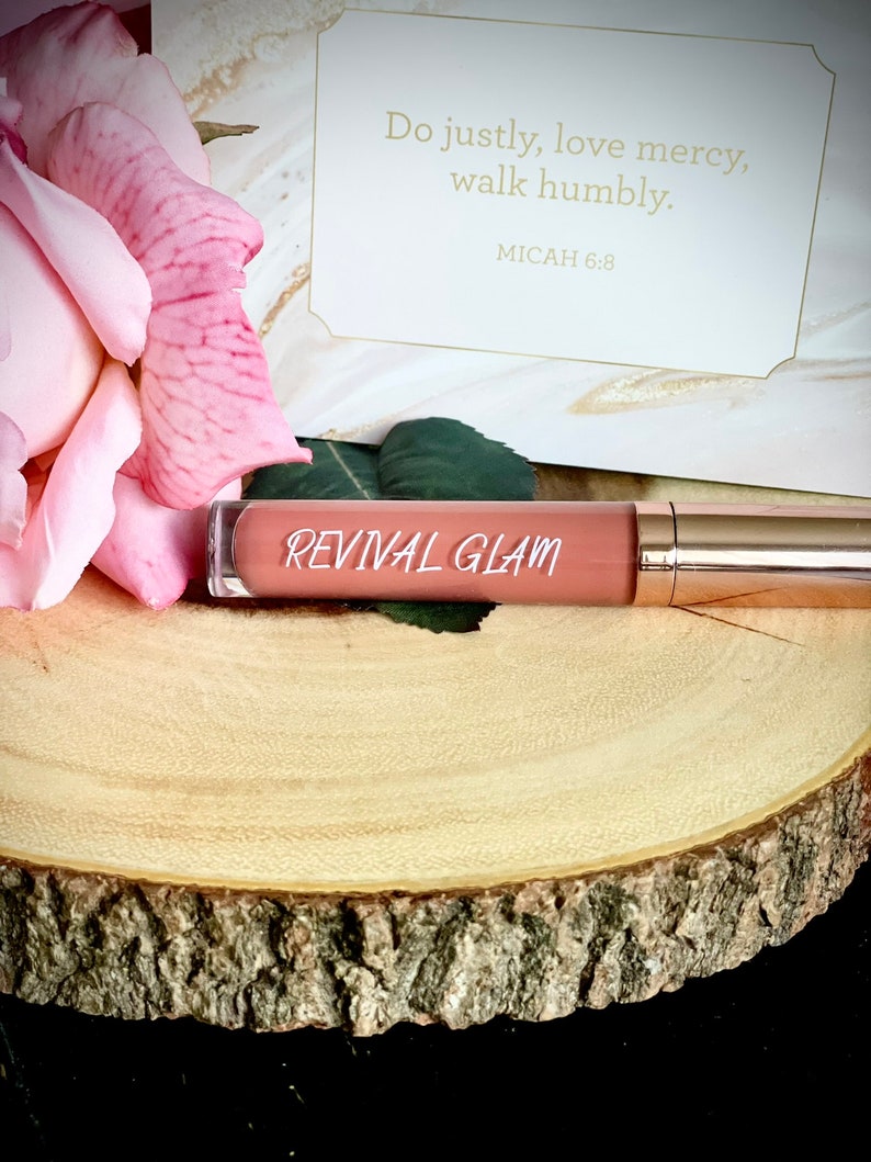 May include: A pink and nude lip gloss tube with the words "REVIVAL GLAM" printed on the label. The tube is sitting on a wooden surface.