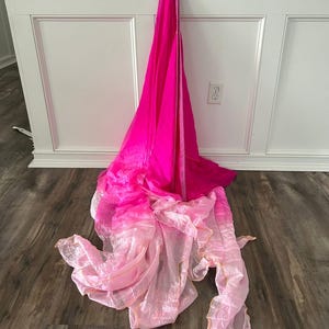 May include: A vibrant, flowing fabric in shades of pink and light pink, draped against a white wall and wooden floor. The fabric appears to be layered, with a gradient effect from deep pink to a lighter, almost translucent pink.