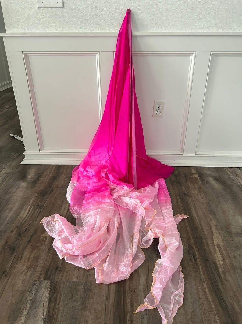 May include: A vibrant, flowing fabric in shades of pink and light pink, draped against a white wall and wooden floor. The fabric appears to be layered, with a gradient effect from deep pink to a lighter, almost translucent pink.