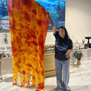May include: A large, flowing fabric in shades of yellow and red, resembling flames, is held up. The fabric has a tie-dye or watercolor effect. The person holding the fabric is wearing a black t-shirt and blue jeans.