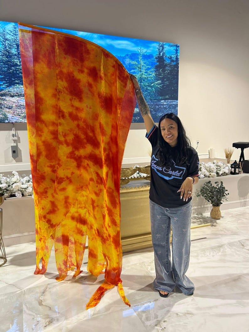 May include: A large, flowing fabric in shades of yellow and red, resembling flames, is held up. The fabric has a tie-dye or watercolor effect. The person holding the fabric is wearing a black t-shirt and blue jeans.