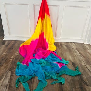 May include: A colorful, flowing scarf with a gradient design. The scarf transitions from red to yellow, then pink, and finally to teal and green. The fabric is draped on a wooden floor.