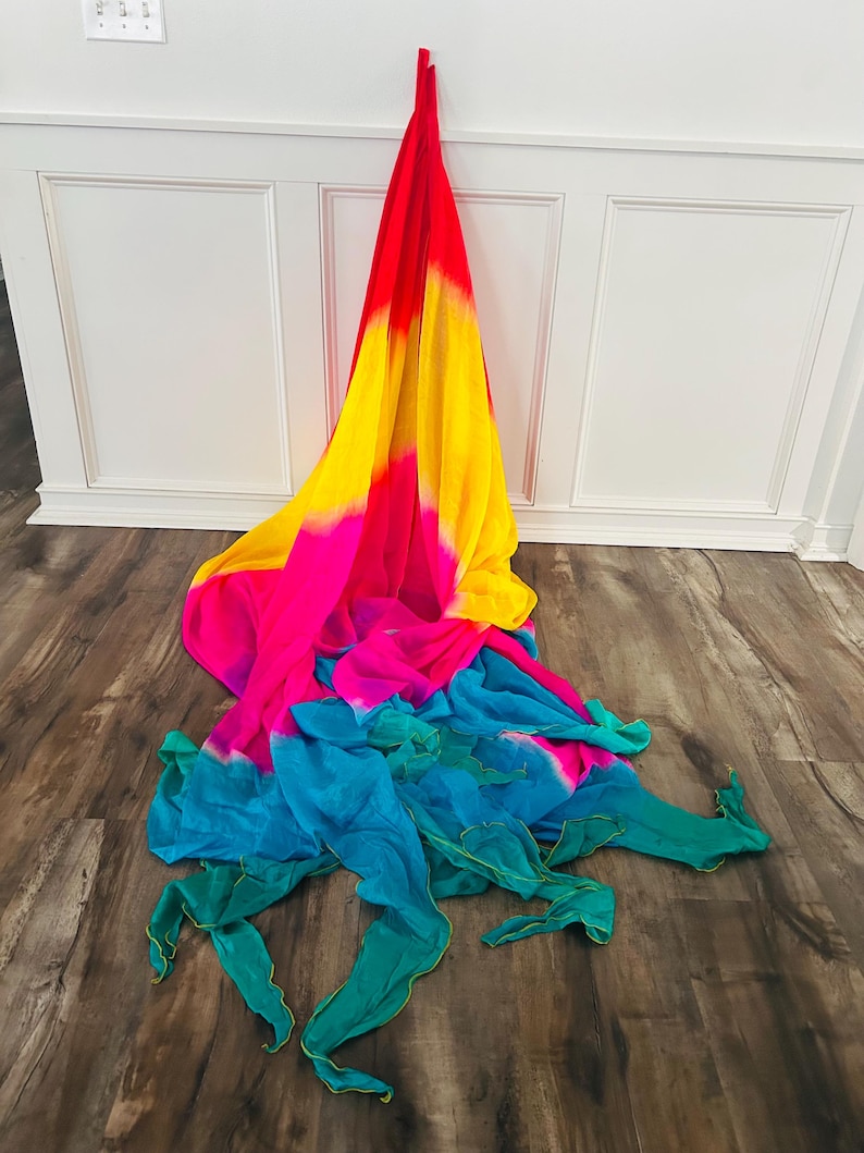 May include: A colorful, flowing scarf with a gradient design. The scarf transitions from red to yellow, then pink, and finally to teal and green. The fabric is draped on a wooden floor.