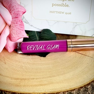 May include: A pink and purple lip gloss tube with the words "Revival Glam" printed on the label. The tube is sitting on a wooden surface.