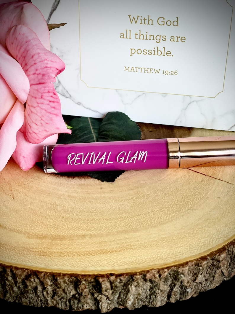 May include: A pink and purple lip gloss tube with the words "Revival Glam" printed on the label. The tube is sitting on a wooden surface.