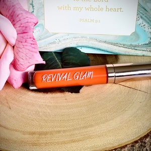 May include: A small orange tube of lip gloss with the words "REVIVAL GLAM" printed on the label. The tube is sitting on a wooden surface with a pink rose in the background. The image also includes a card with the text "I will give thanks to the Lord with my whole heart. PSALM 9:1"