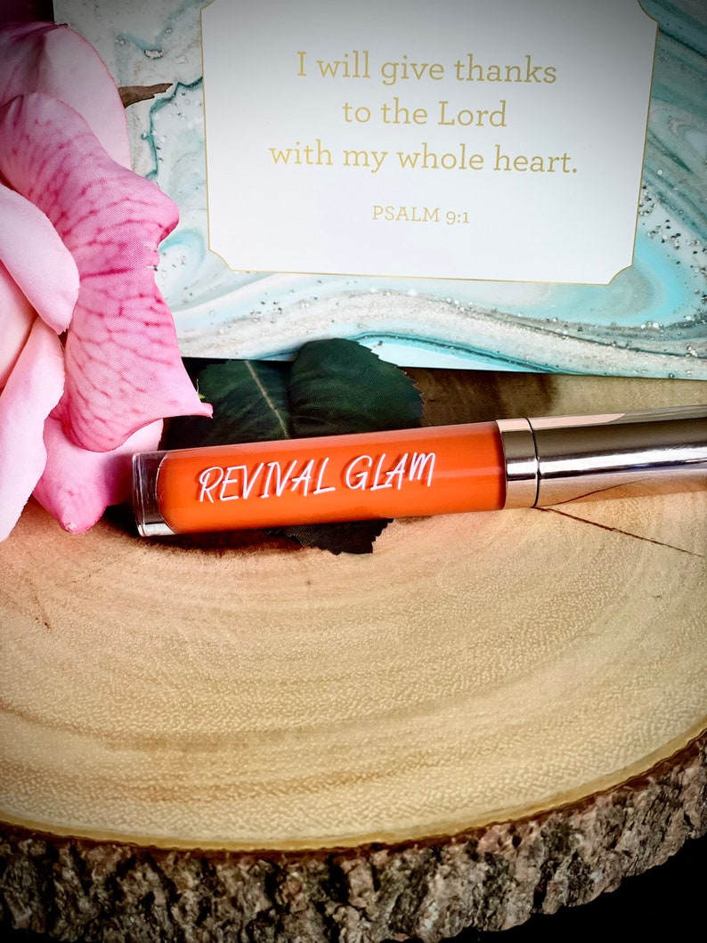 May include: A small orange tube of lip gloss with the words "REVIVAL GLAM" printed on the label. The tube is sitting on a wooden surface with a pink rose in the background. The image also includes a card with the text "I will give thanks to the Lord with my whole heart. PSALM 9:1"