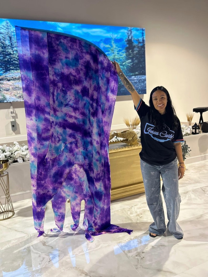 May include: A large, flowing fabric in shades of purple and blue, with a tie-dye pattern. The fabric is held up, showcasing its length and design. The image also shows a person wearing a black t-shirt with the text "Jesus Cartel" and light blue jeans.
