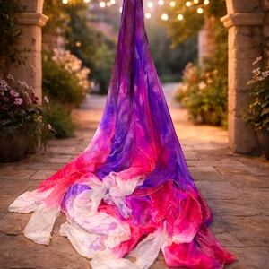 May include: A flowing, vibrant fabric in shades of purple, pink, and white, draped on a stone walkway. The fabric's colors blend together, creating a watercolor effect. The scene is set outdoors with a blurred background of greenery and soft lighting.