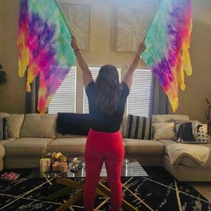 May include: A person holding two colorful, tie-dye flags with shades of purple, pink, yellow, and green. The flags are held up in front of a window. The person is wearing a black top and red pants.