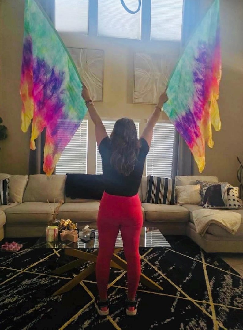 May include: A person holding two colorful, tie-dye flags with shades of purple, pink, yellow, and green. The flags are held up in front of a window. The person is wearing a black top and red pants.