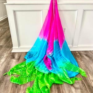 May include: A vibrant, multi-colored scarf with a gradient design. The scarf transitions from bright pink to turquoise blue, and then to a bright green. The scarf is draped against a white wall and wooden floor.