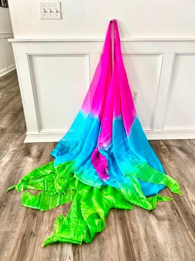 May include: A vibrant, multi-colored scarf with a gradient design. The scarf transitions from bright pink to turquoise blue, and then to a bright green. The scarf is draped against a white wall and wooden floor.