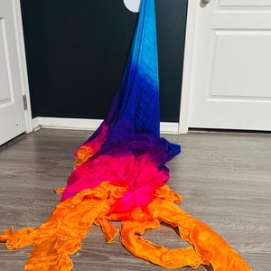 May include: A colorful, flowing fabric in shades of blue, purple, pink, and orange. The fabric is draped on a wooden floor, with the blue end hanging against a dark wall. The colors blend in a gradient effect, creating a vibrant and dynamic visual.