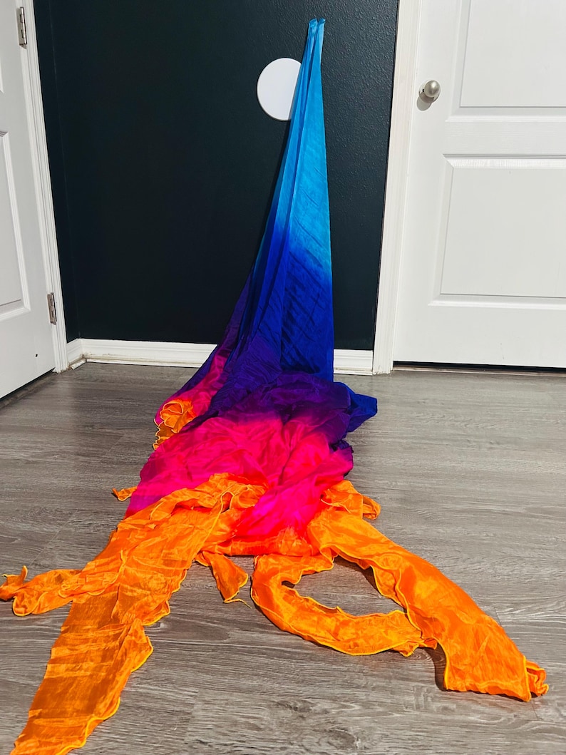 May include: A colorful, flowing fabric in shades of blue, purple, pink, and orange. The fabric is draped on a wooden floor, with the blue end hanging against a dark wall. The colors blend in a gradient effect, creating a vibrant and dynamic visual.