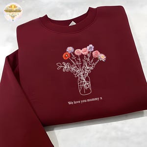 Peut inclure: Sweat-shirt bordeaux avec un motif de bouquet floral. Le bouquet, dans un vase, présente des fleurs roses, violettes et orange. Le texte "We love you mummy x" est imprimé en dessous.